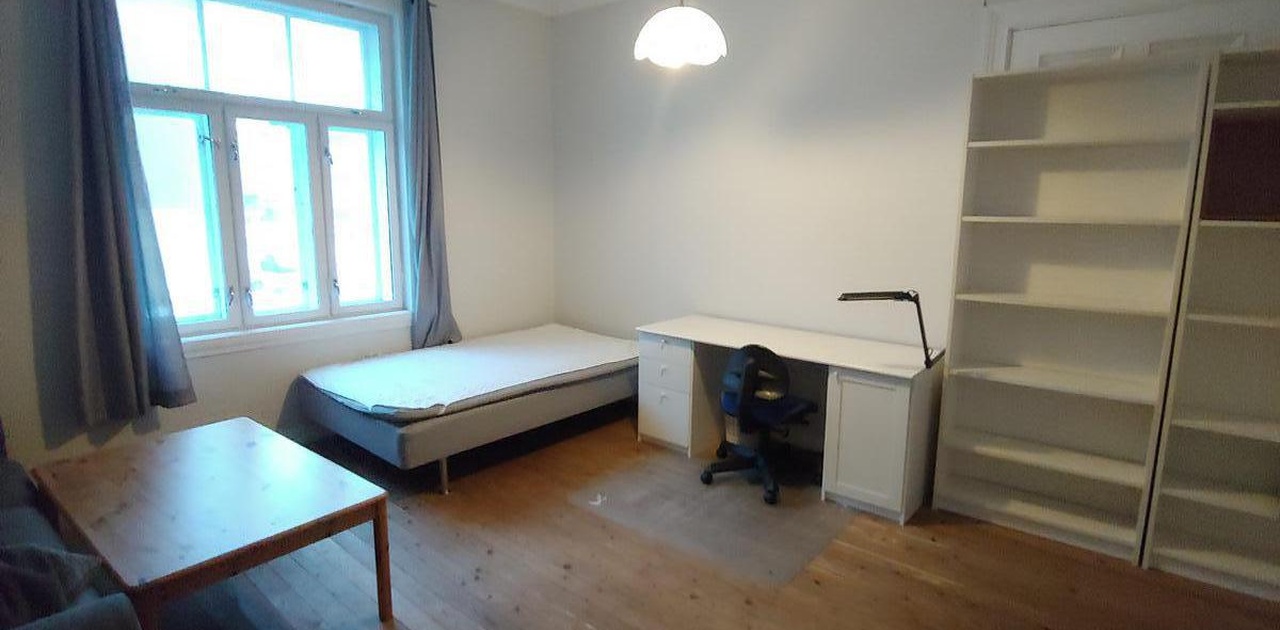Hybel bilde mot vindu. Viser bord, seng, pult og bokhylle.