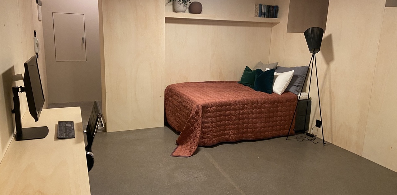 Bilde tatt fra kjøkken, mot seng/sofa. Plassbygget hylle over senga med plass til dekorasjon, bøker m.m. Sengen har oppbevaring under.
