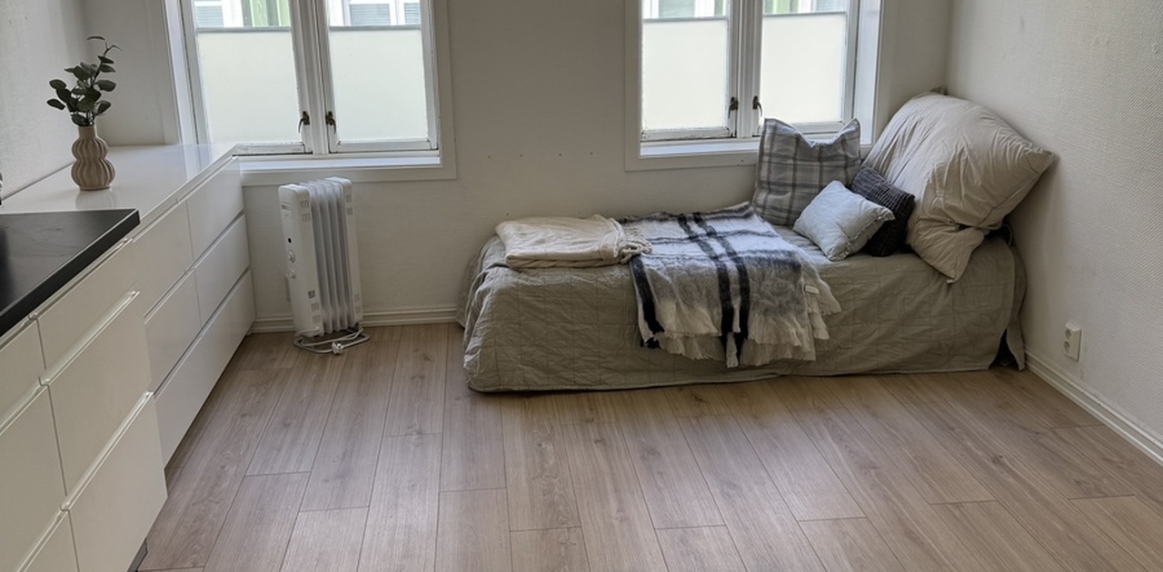 Rommet er lyst og romslig, med store vinduer som slipper inn godt med naturlig lys. Det har lyse tregulv og hvitmalte vegger som gir en moderne og ren følelse. Eget kjøkken er integrert i rommet, med hvite skap, sort benkeplate, kjøleskap og vask