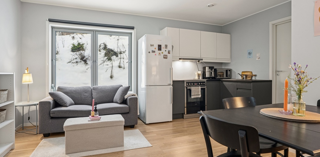 Lys stue og åpen kjøkken - moblert med sofaen, spisebordet med stoler, hylla, og kjøleskap.