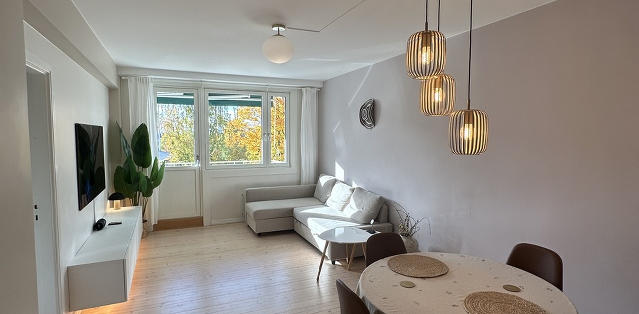 Stuen er innredet med et lyst tregulv, hjørnesofa og spiseplass opplyst av moderne pendellamper.