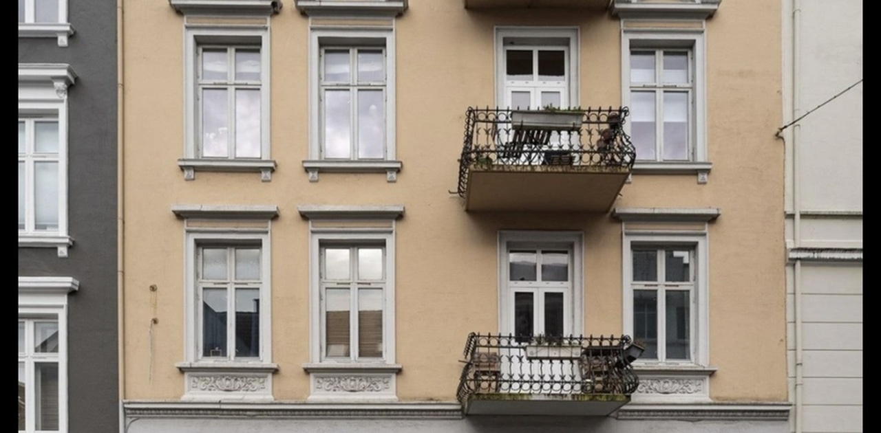 Bygårdens gule murgårdsfasade har smijernsbalkonger og detaljrike vindusomramminger mot en brolagt gate.