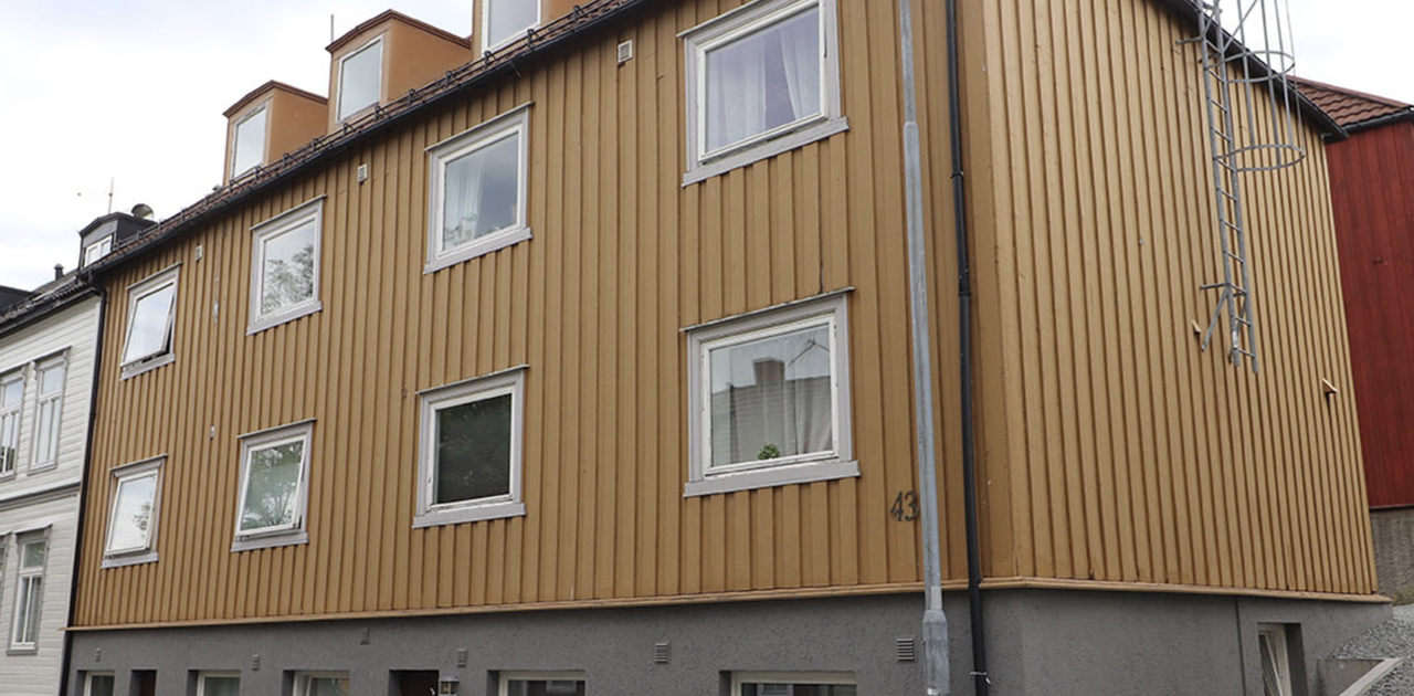 Bygningen har stående, brun/gul trepanel med grå pusset underetasje og store vindusflater mot gaten.