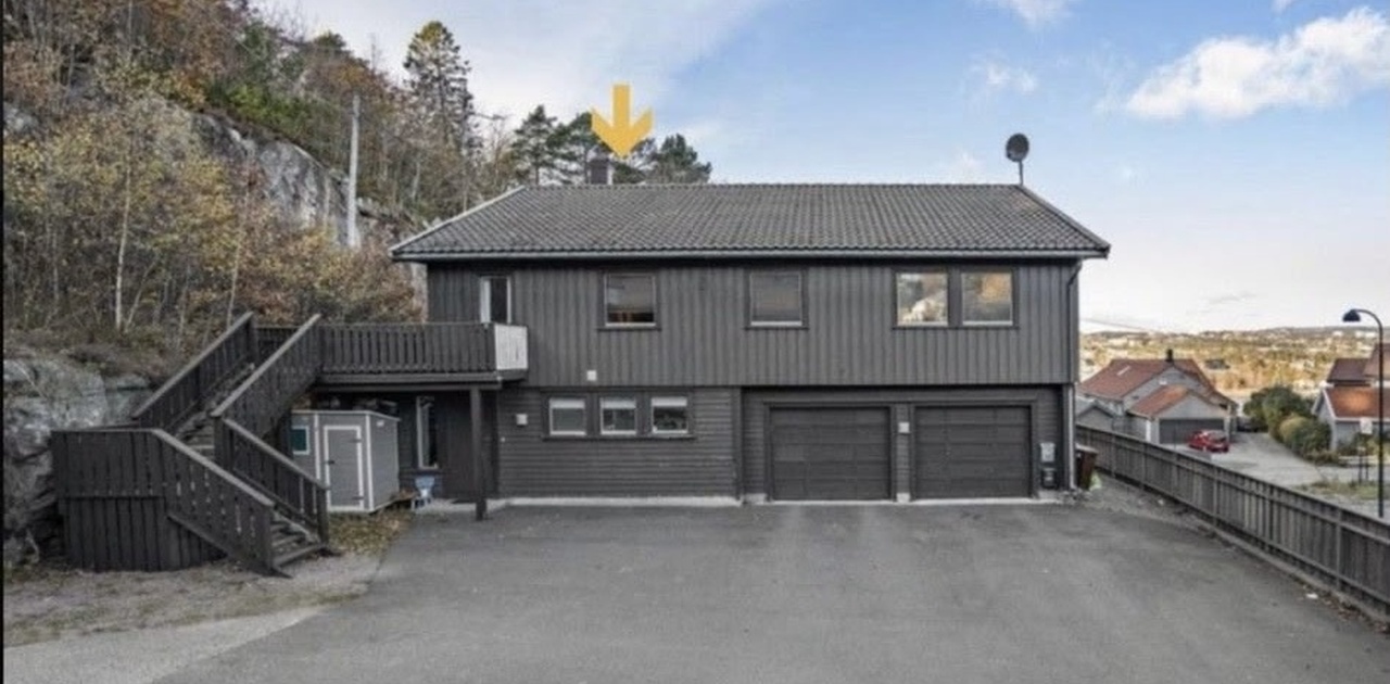 Bygningen har mørk trepanel, integrert dobbelgarasje og en stor asfaltert gårdsplass med utvendig trapp til balkongen.