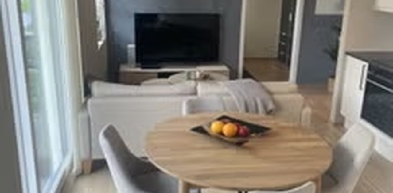 Stuen har en åpen løsning med lyst parkettgulv, rund spiseplass og en mørk kontrastvegg bak TV-kroken.