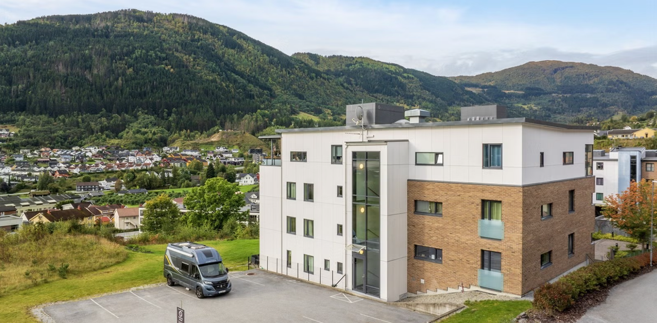 Bygget har en moderne fasade i kombinert platekledning og teglstein, med tilhørende parkeringsplass og mektige fjell i bakgrunnen.
