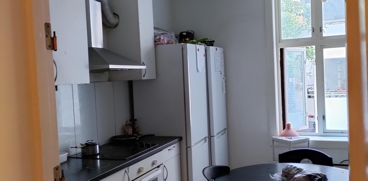 Gjennom en hvelvet døråpning ses kjøkkenet med hvite skapfronter, to store kombiskap og en rund spiseplass ved vinduet.
