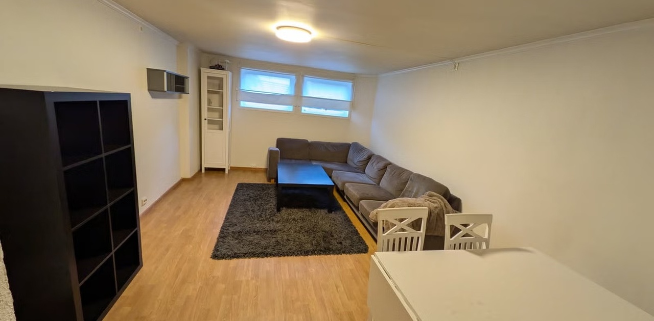 Stuen har parkettlignende gulv og er innredet med en stor grå hjørnesofa, salongbord, en hvit reol og en spisegruppe.