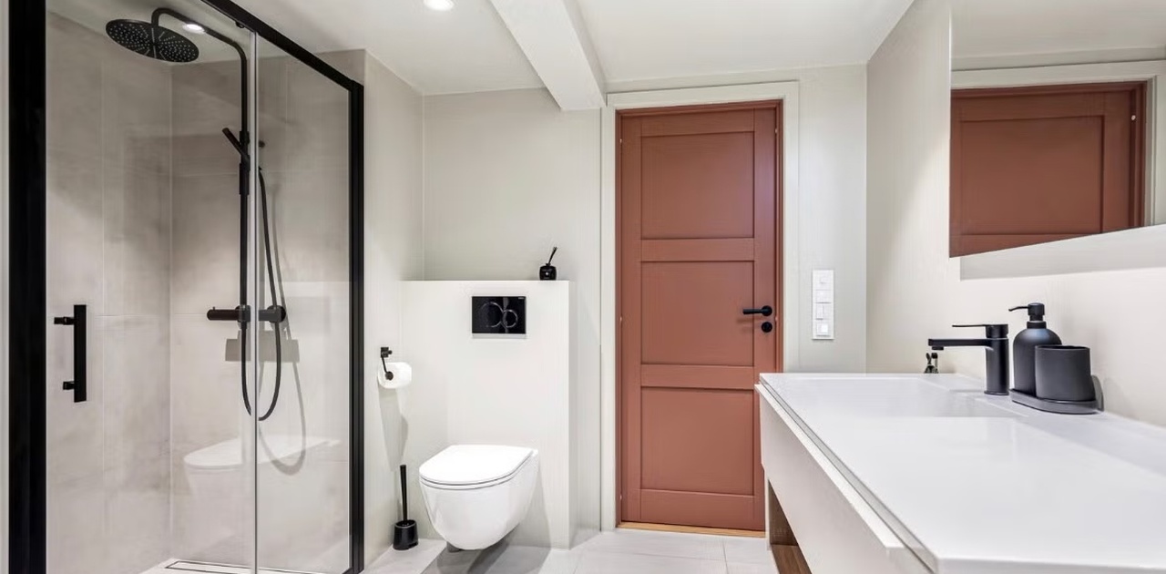 Svarte detaljer og en karakteristisk rødbrun dør gir fin kontrast til det moderne badet med glassdusj og vegghengt toalett.