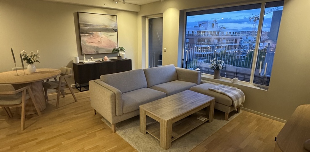 Stuen har parkettgulv, spiseplass og en lys sofagruppe plassert ved et stort vindu med utsyn over byen.