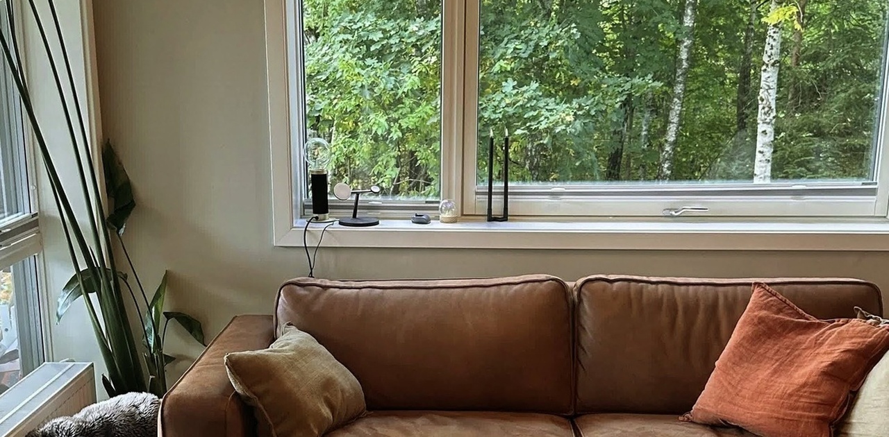Stuen har en brun skinnsofa plassert under et stort vindu med utsikt mot grønn skog.