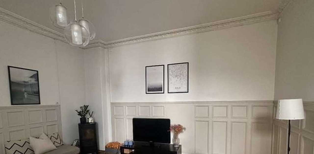 Klassiske brystningspaneler og takstukkatur rammer inn stuen, som er innredet med en stor hjørnesofa, peisovn og parkettgulv.