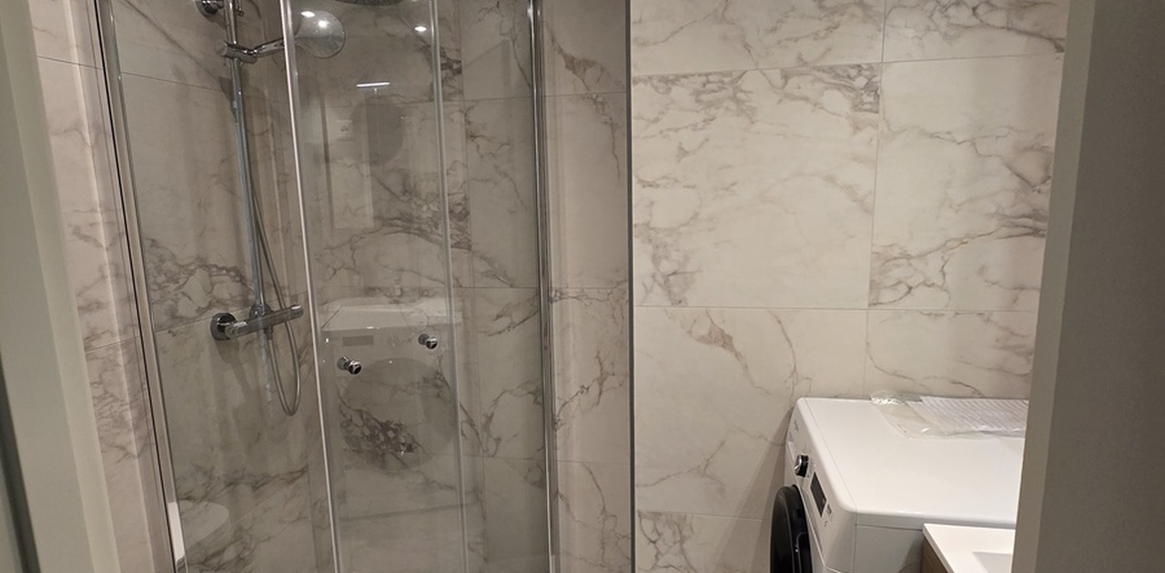Marmormønstrede fliser dekker gulv og vegger på det moderne badet, som har glassdusj, vegghengt toalett og vaskemaskin.