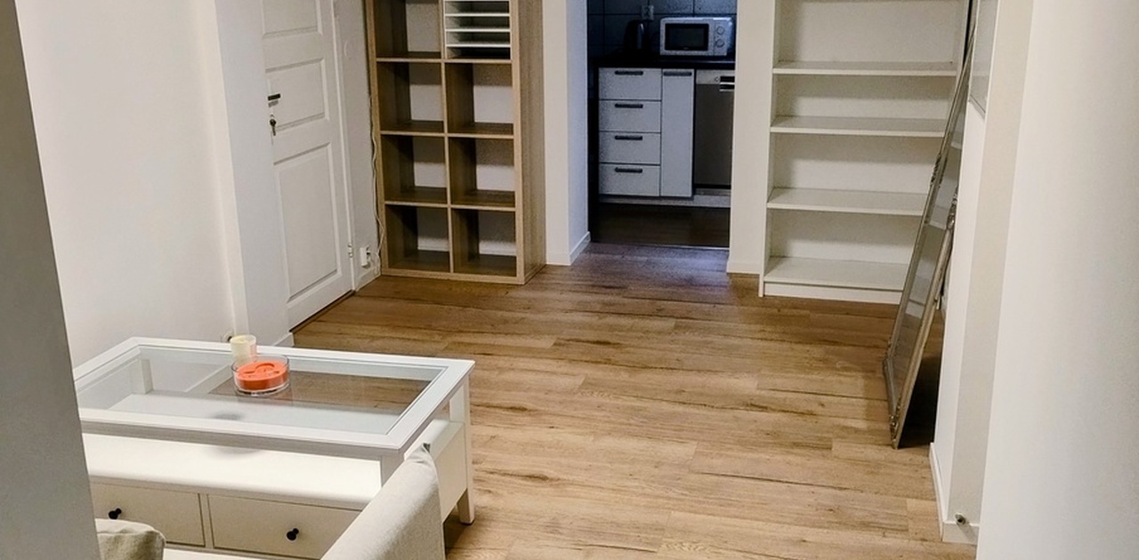 Stueområdet har tregulv-mønstret laminat og praktiske oppbevaringshyller, med kikk inn mot det tilstøtende kjøkkenet.