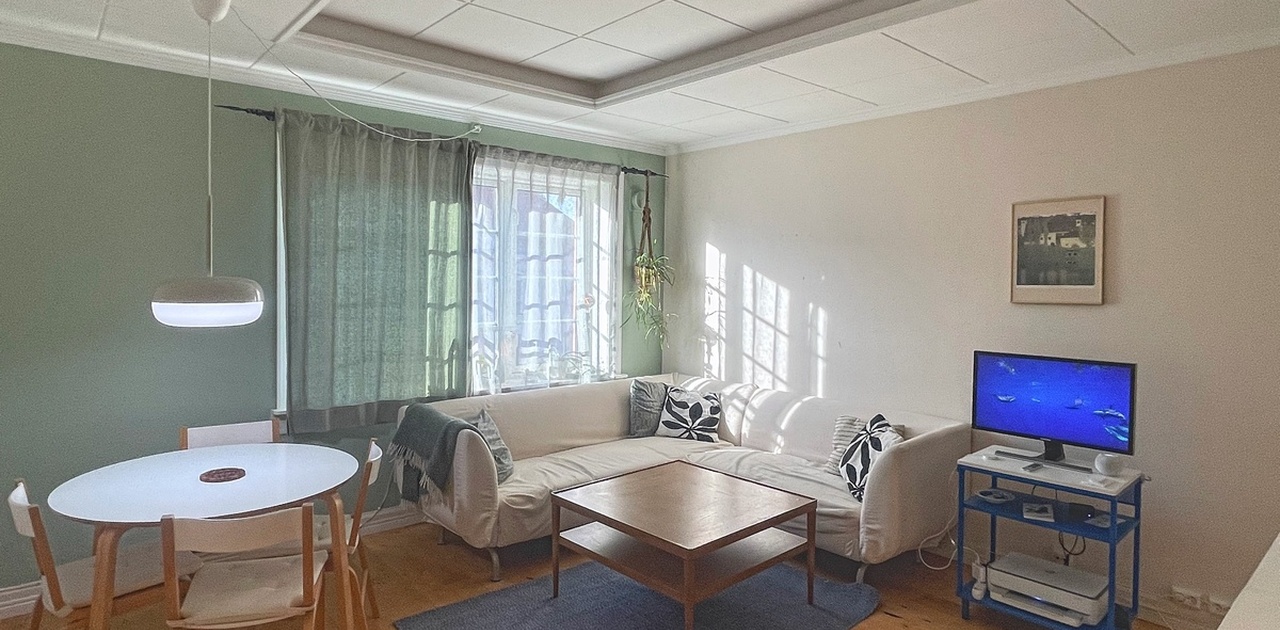 Sollys strømmer inn i stuen, som er innredet med en hvit hjørnesofa, salongbord og en spiseplass ved den grønne fondveggen.