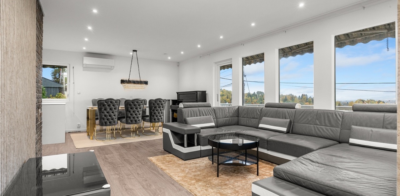Stuen byr på en stor hjørnesofa i skinn, spiseplass med hengt lysekrone, integrerte downlights i taket og store vindusflater med vidstrakt utsikt.