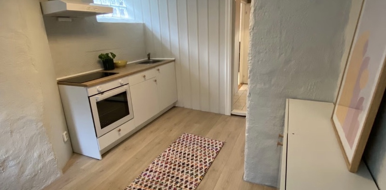 Kjøkkenkroken har hvit innredning med integrert stekeovn og platetopp, kombinert med hvitpanelte vegger og lyst laminatgulv.