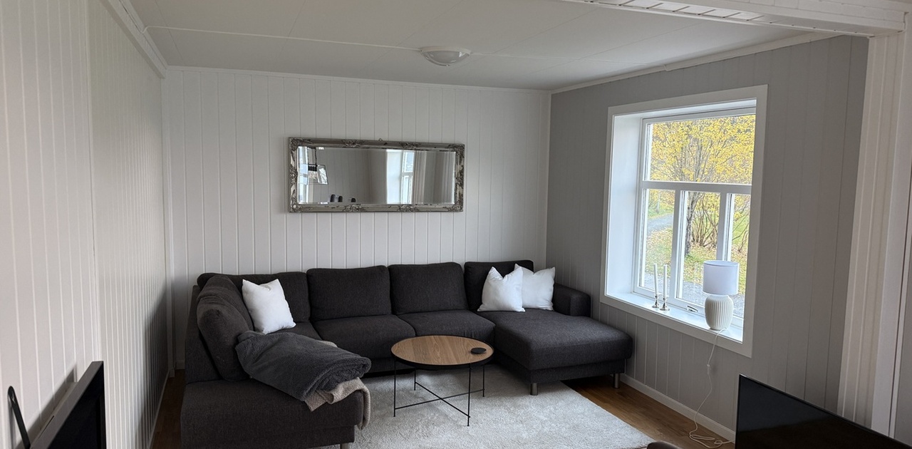 En stor mørk U-sofa utgjør sittegruppen i stuen, som har parkettgulv, hvitmalte panelvegger og et vindu med utsikt mot skjermede omgivelser.