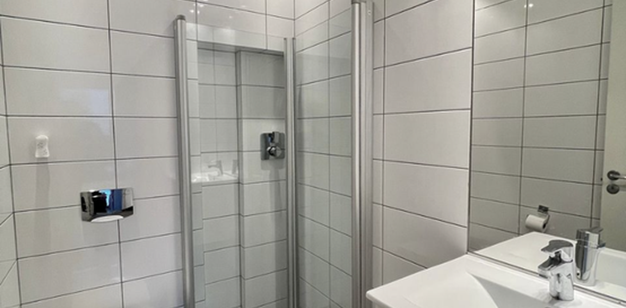 Badet har stilrene hvite veggfliser, mørkegrå gulvfliser, glassinnbygd dusjhjørne med takdusj og vegghengt toalett.