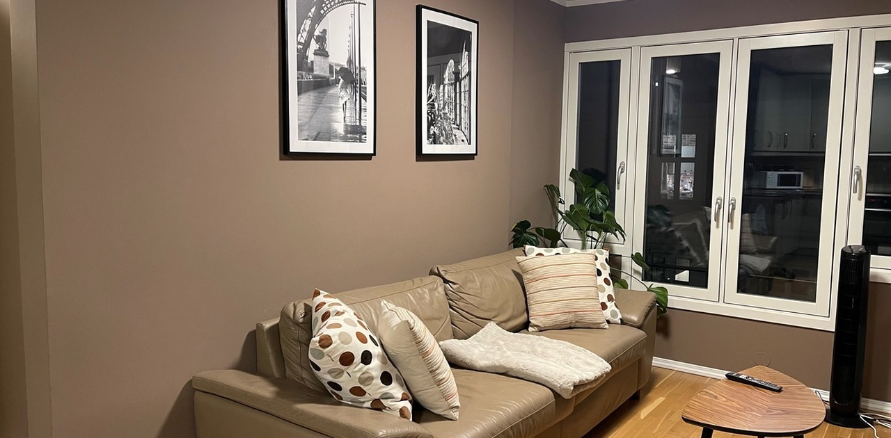 En beige skinnsofa med dekorerte puter står plassert mot en lun brun vegg prydet med svarthvitt-bilder.