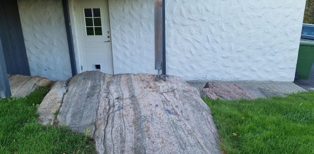 Inngangspartiet har hvitpussa fasade og hvit ytterdør, rammet inn av et markant naturlig svaberg i gressplenen.