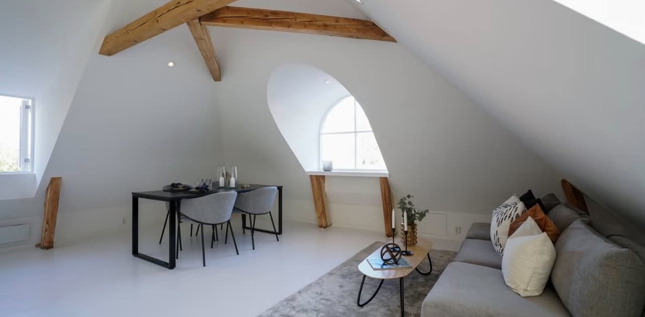Synlige trebjelker og et karakteristisk buevindu gir særpreg til denne stilrene loftstuen innredet med sofa og spiseplass.