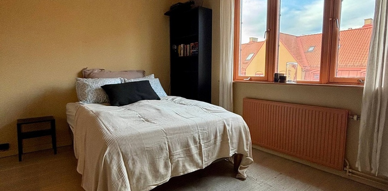 Leiligheten har varme gule vegger, lyst eike gulv og et tredelt vindu som gir utsyn mot nærliggende tak. Det er også radiator.