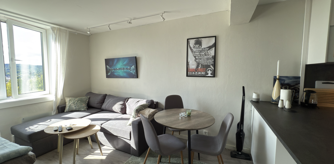 Stuen er innredet med en grå sofa, rundt spisebord og et stort vindu som slepper inn rikelig med dagslys og gir utsikt.