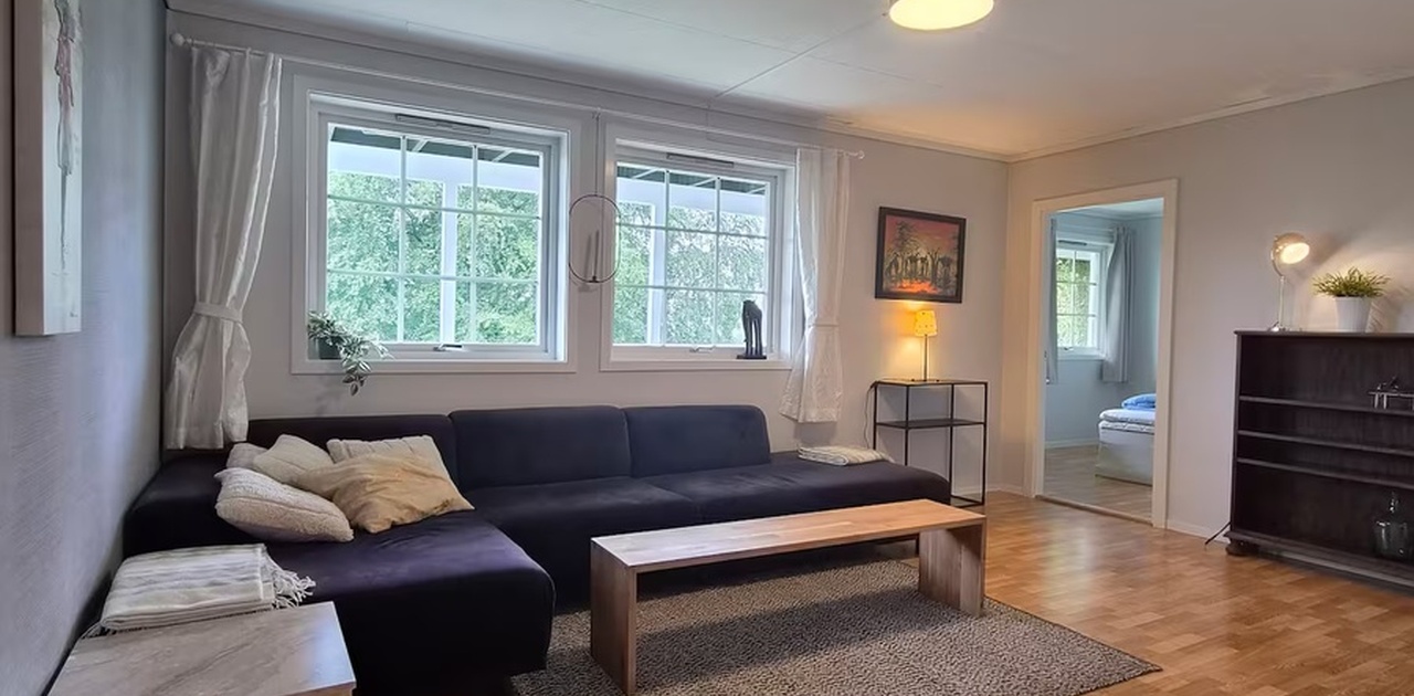 Stuen er innredet med en stor, mørk hjørnesofa, et enkelt trebord på gulvteppe og parkettmønstret gulv, med lysinnslipp fra sprossede vinduer.