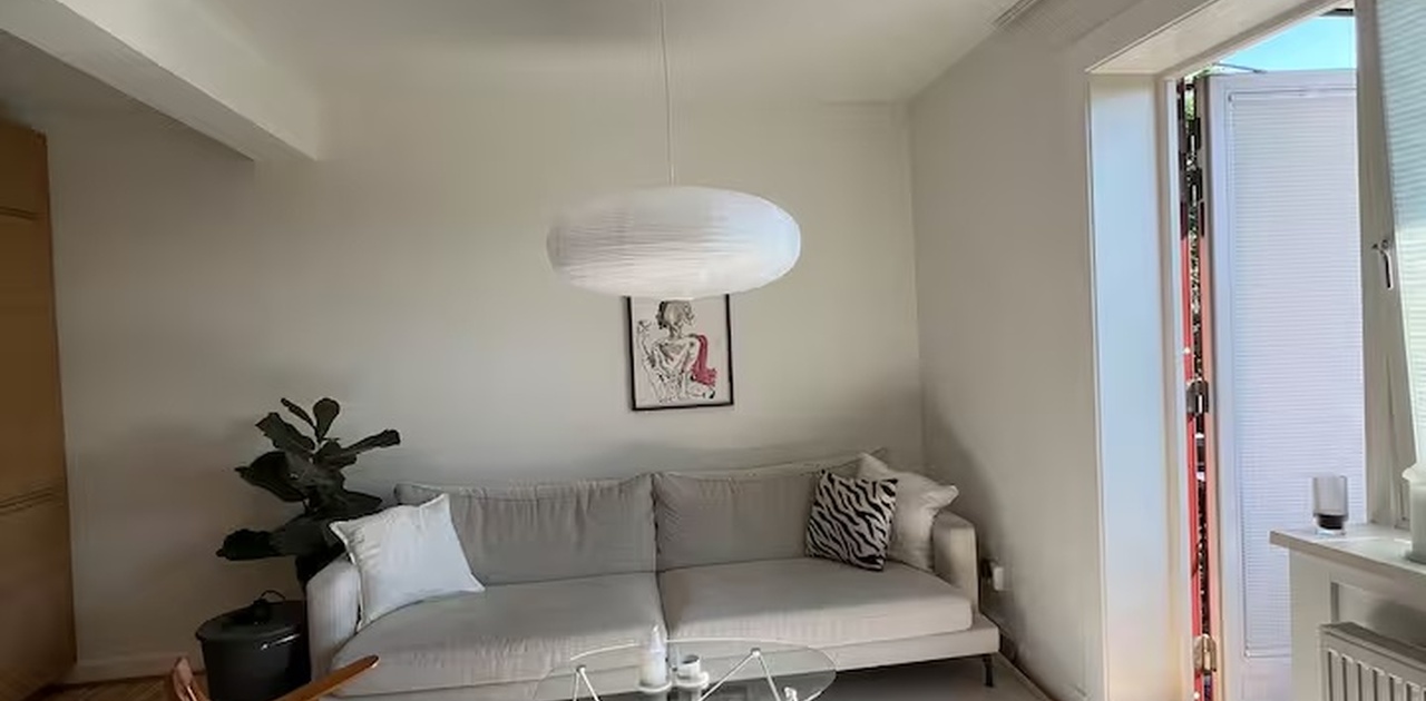Stuen har en lys sofa, glassbord og retro lenkestol i tre, med et stort teppe og utgang til balkong.