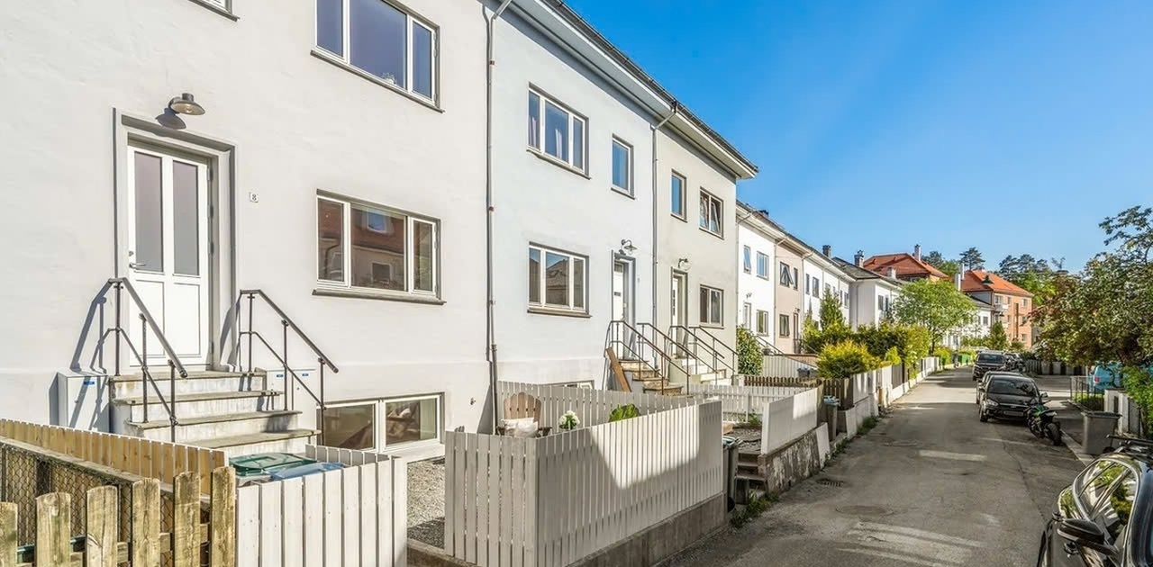 Hvite murfasader med trapper opp til inngangspartiene ligger på rekke langs en solfylt boliggate.