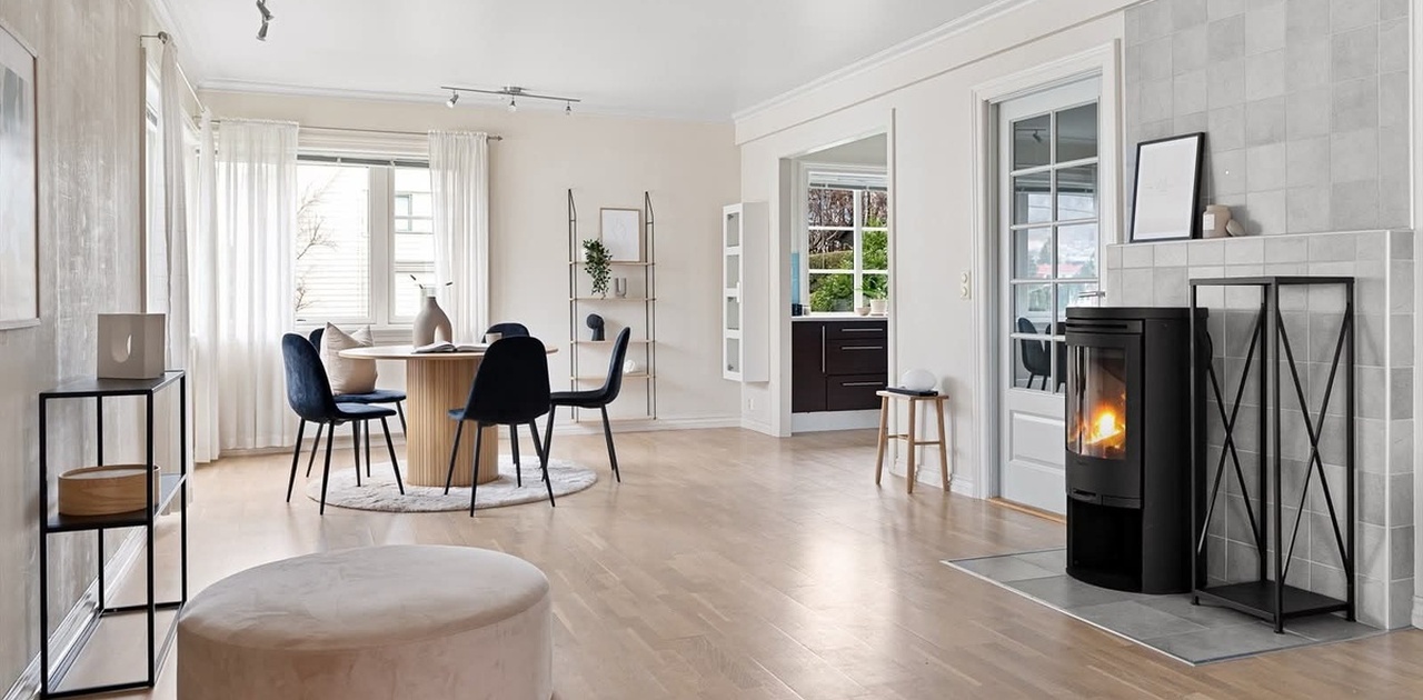 En stilig peisovn skaper stemning i den åpne stuen med lyst parkettgulv og spiseplass ved vinduet.