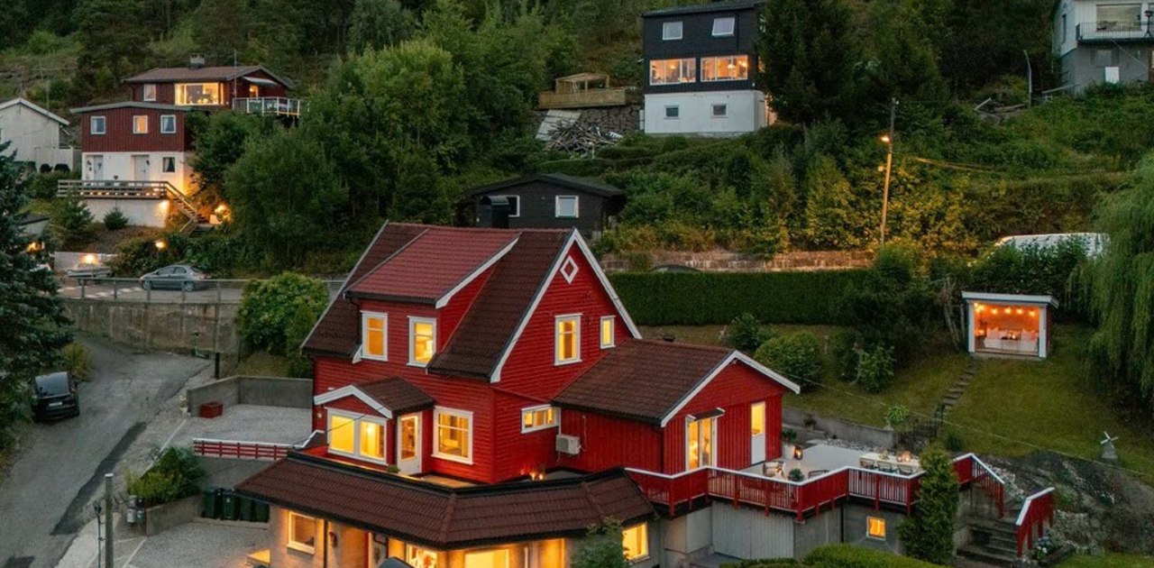 Det rødmalte huset ligger lunt til i et kupert terreng med tett skog i bakkant, omgitt av en stor terrasse og opplyste vinduer i skumringen.
