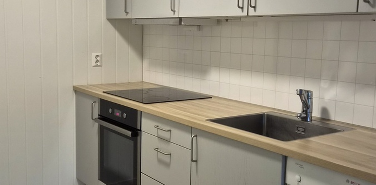Kjøkkenet har grå fronter, lys benkeplate og hvitevarer som integrert stekeovn, platetopp og oppvaskmaskin.