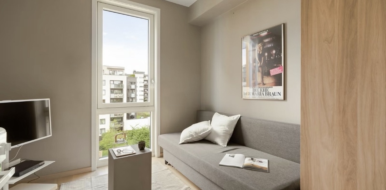Oppholdsrommet er innredet i dempede beige- og gråtoner med en kombinert sovesofa, TV-løsning og et stort vindu som gir rikelig med dagslys.