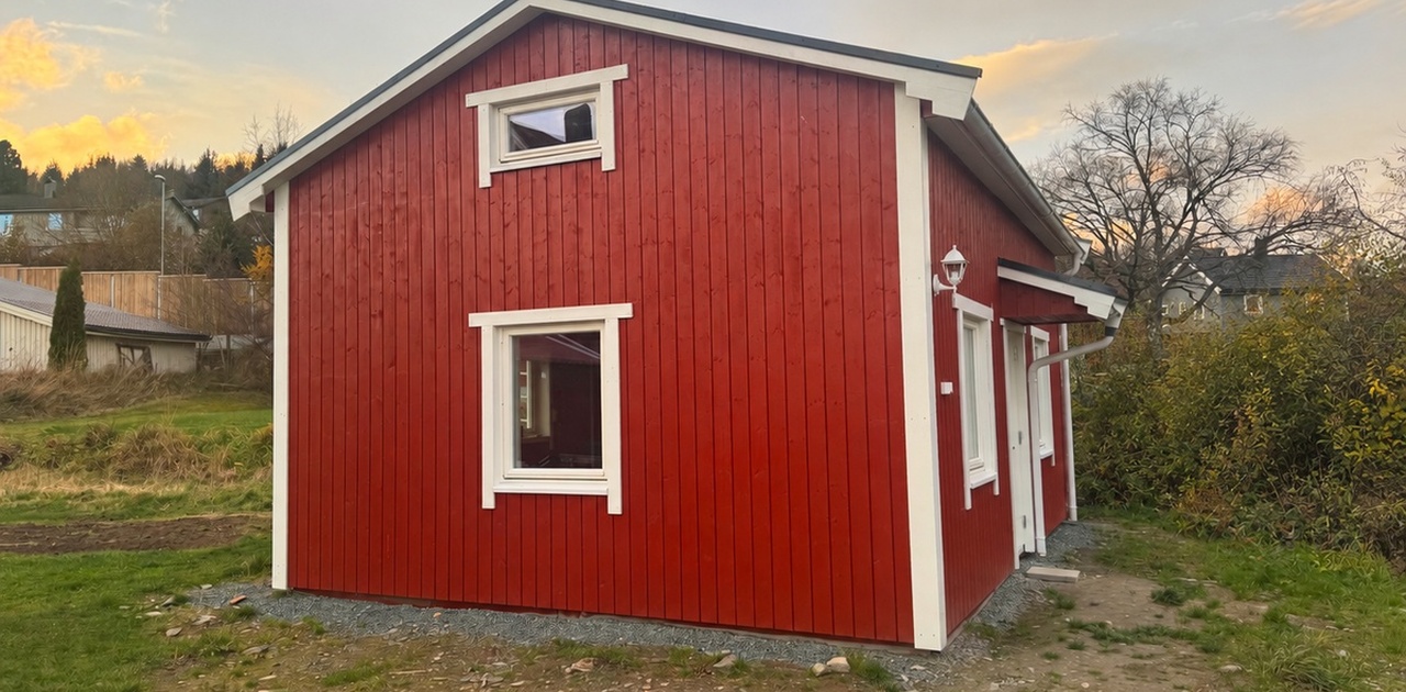 Bygningen har stående rød trepanelt kledning med hvite vindusrammer og omkranses av et gresskledd uteområde.