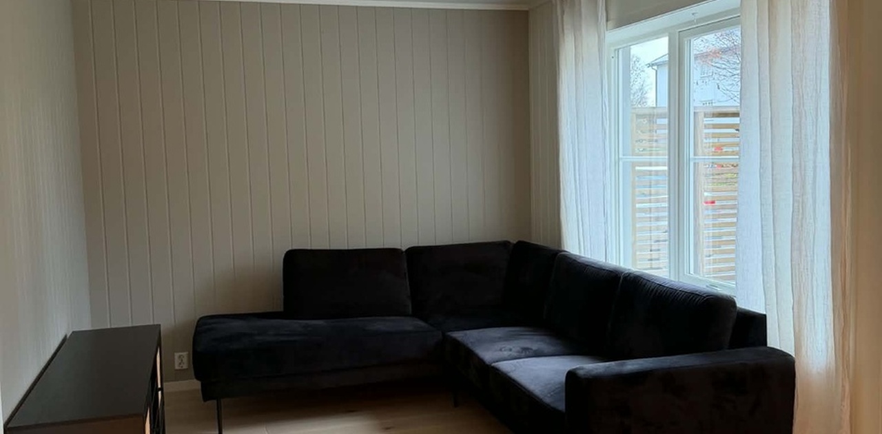 Stuen har lys parkett på gulvet, malt panel på veggene og en mørk hjørnesofa ved vinduet.