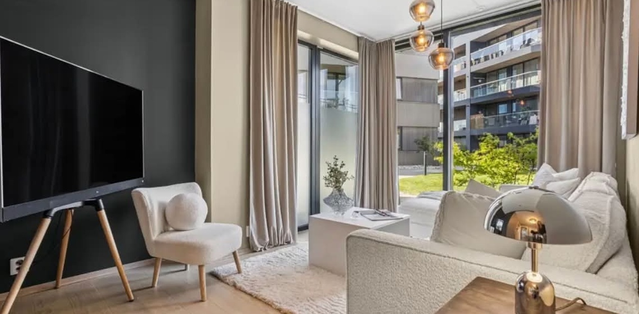Stuen er innredet med en lys sofa, hengende glasslamper og en TV på trefot mot en mørk kontrastvegg ved store vindusflater.