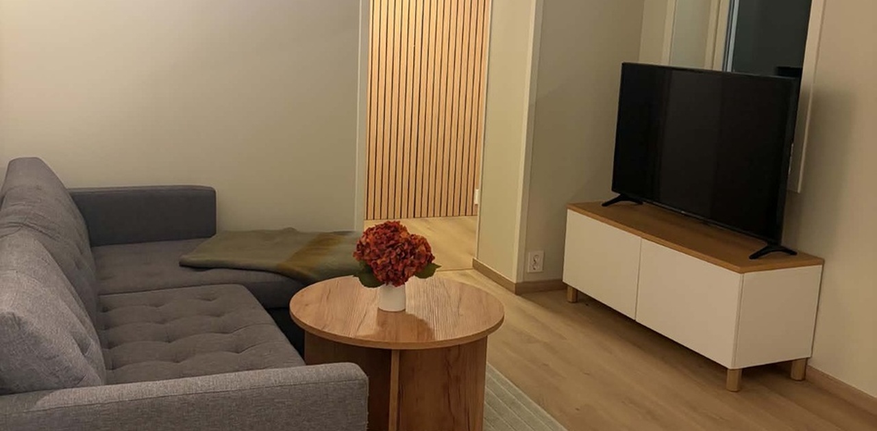Stuen har en grå hjørnesofa og rund salongbord i tre, med innsyn til gangen med moderne spilevegg.