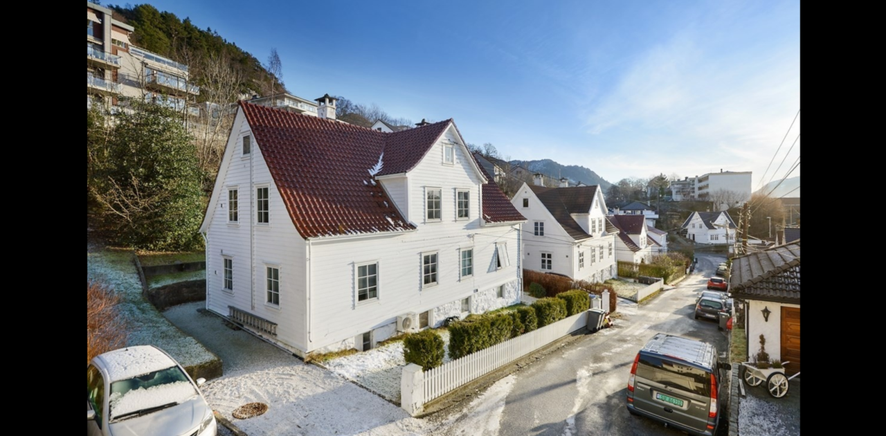 Bygningen er et klassisk hvitmalt trehus med rødt takstein, omkranset av en hekk og plassert i en rolig gate ved foten av en skogkledd ås.