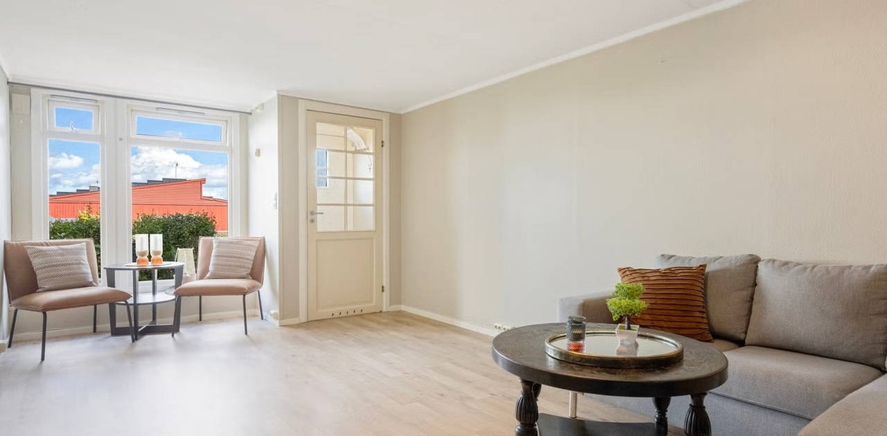 Store vinduer slipper godt med dagslys inn i stuen, som er innredet med en grå sofa og en koselig sittegruppe ved vinduet.