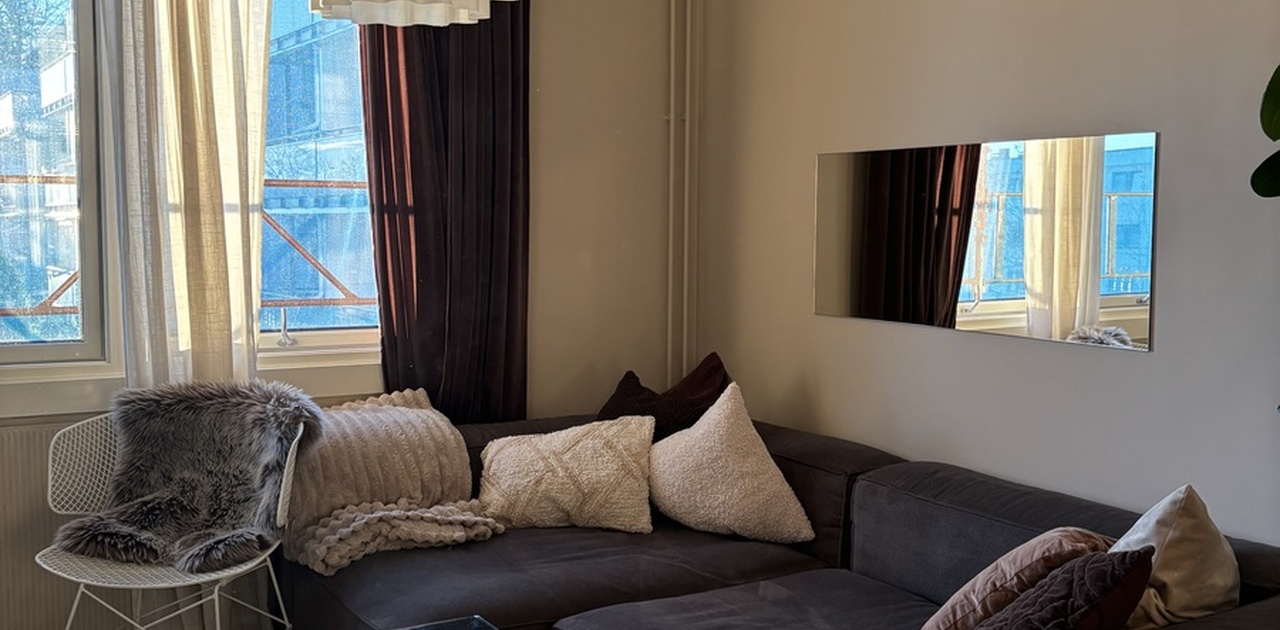 Stuen har en mørk hjørnesofa med rikelig med pynteputer, kombinert med et gjennomsiktig glassbord og en iøynefallende pendellampe.