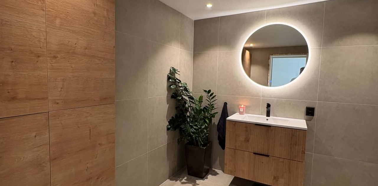 Badet har beige fliser på vegger og gulv, kombinert med et svevende servantmøbel i trestruktur og et rund speil med integrert belysning.
Det er egen inngang samt separert wc