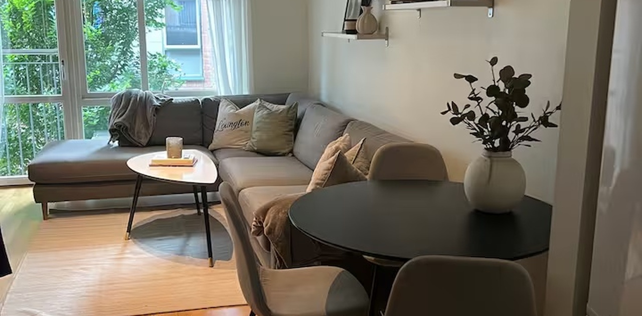 Stuen har parkettgulv, en stor grå hjørnesofa og spiseplass, samt store vindusflater mot balkong og grønne omgivelser.