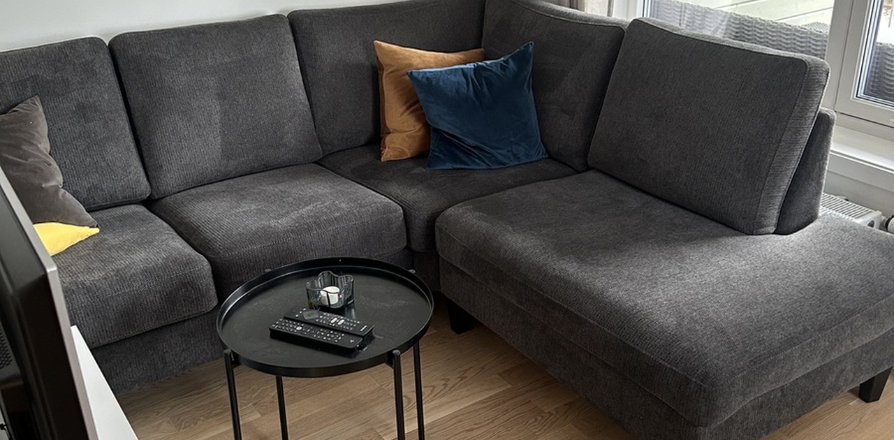 Stuekroken har en mørkegrå hjørnesofa med pynteputer, et lite rundt salongbord og lyst parkettgulv.
