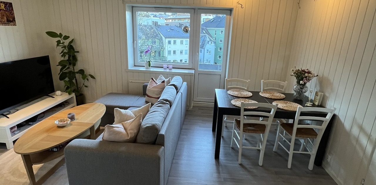Stuen kombinerer en grå hjørnesofa og TV-krok med en spiseplass ved vinduet som vender ut mot bebyggelsen.