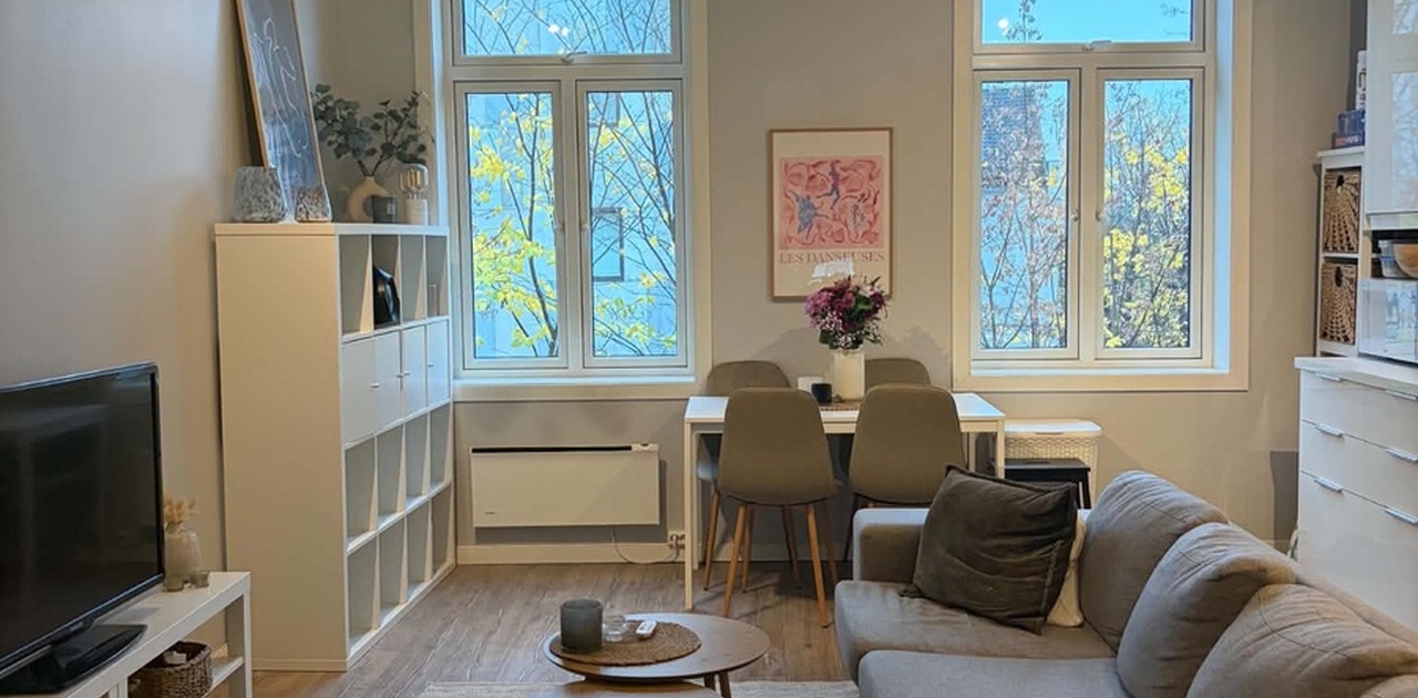 Stuen er innredet med en grå hjørnesofa, runde trebord og en spiseplass plassert foran to høye vinduer. Det er høyt under taket og god beslysning fra vinduene.
