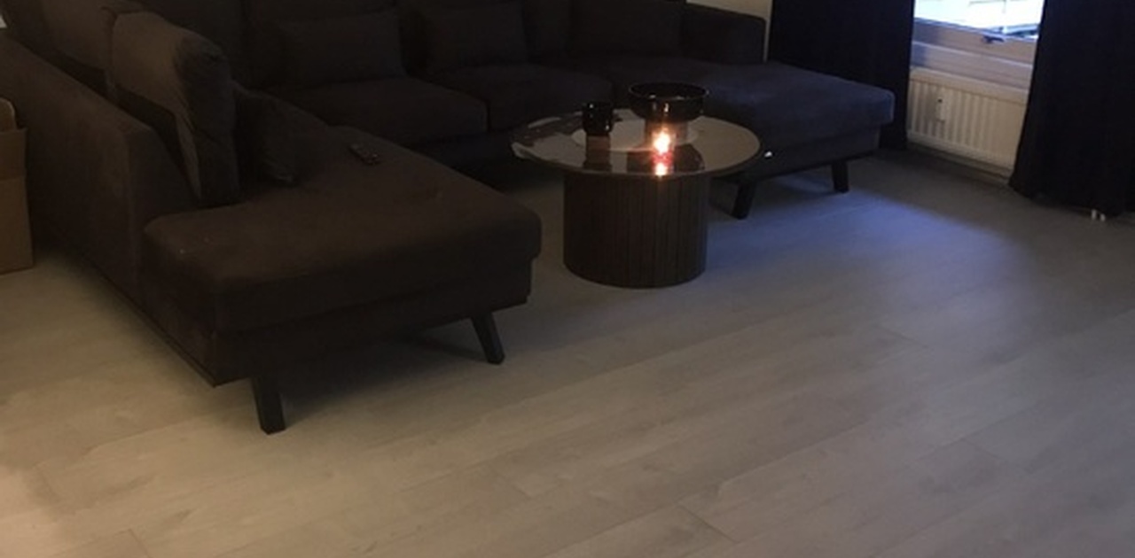 Stuen er innredet med en stor, mørk hjørnesofa og et rundt salongbord mot et lyst laminatgulv.