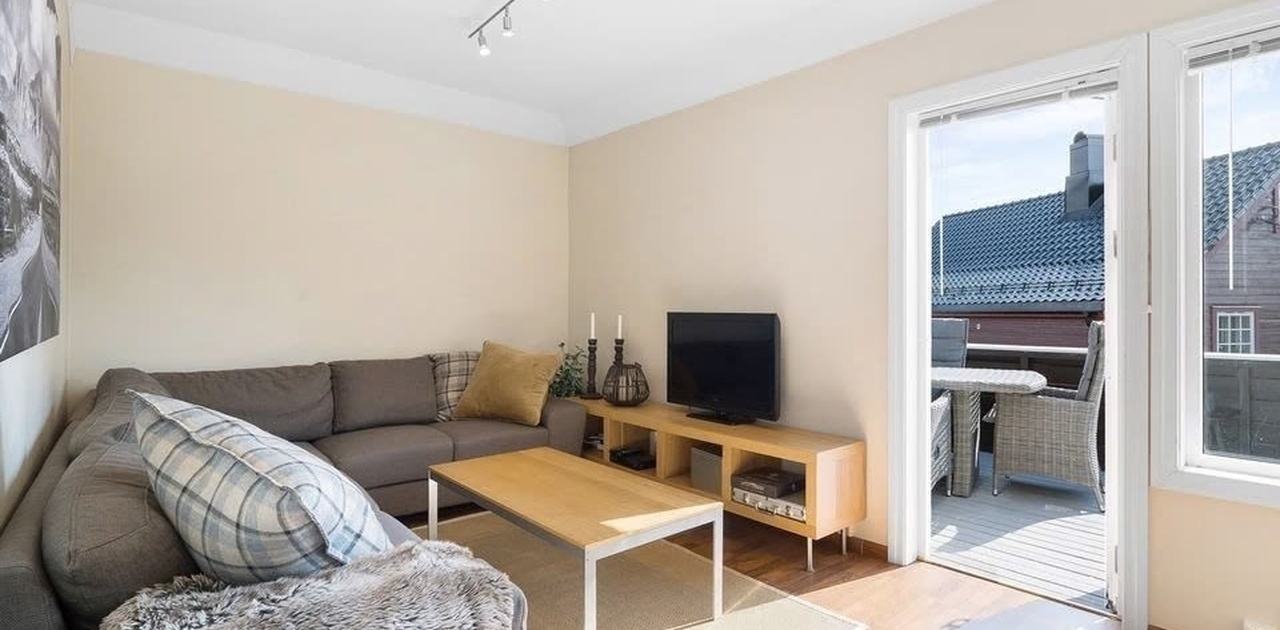 Stuen er innredet med en stor grå hjørnesofa og lav TV-benk, og har direkte utgang til en solrik balkong.