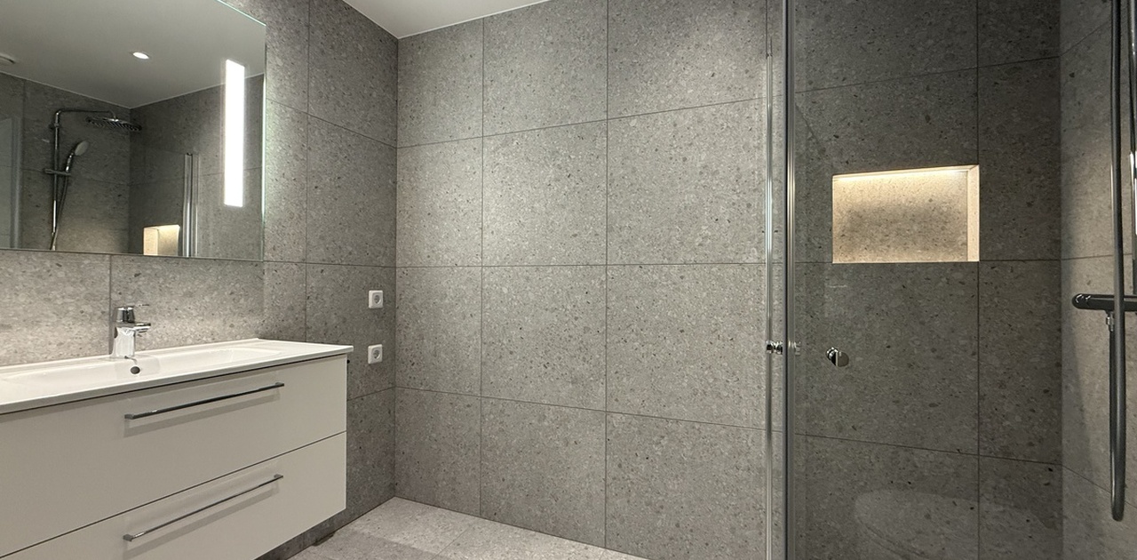 Store, grå fliser og et glassdusjhjørne med takdusj og belyst nisje preger dette moderne badet med hvit servantinnredning.